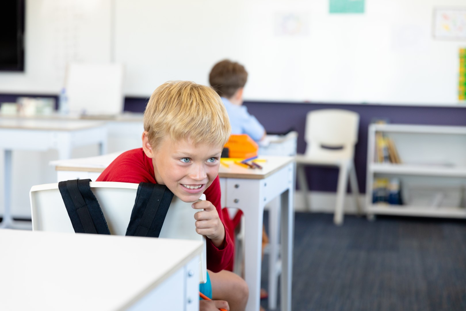 Smiling caucasian elementary schoolboy looking away while leaning on chair in classroom. unaltered, childhood, education and back to school concept.