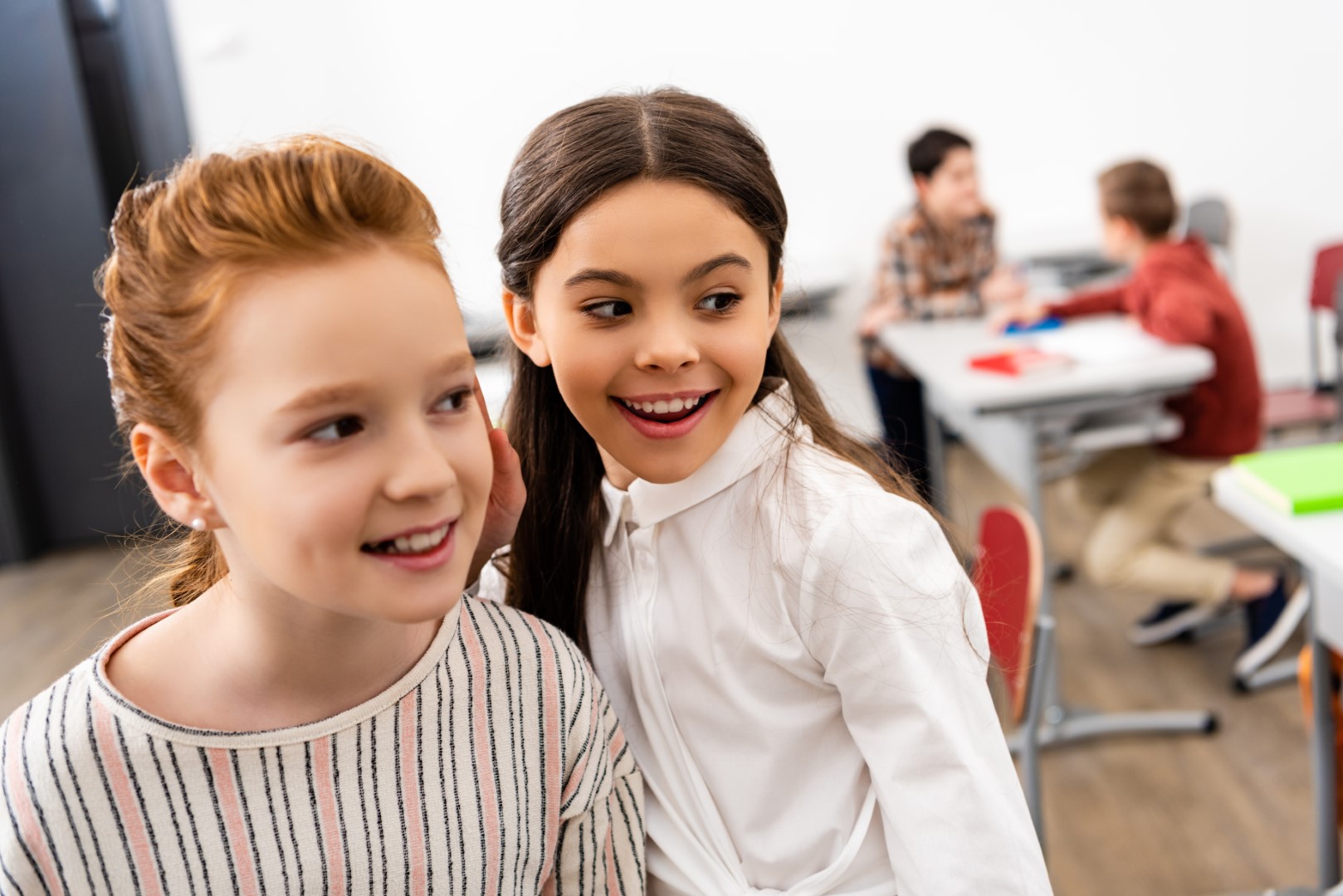 Two cute schoolgirls smiling and looking away in classroom during brake